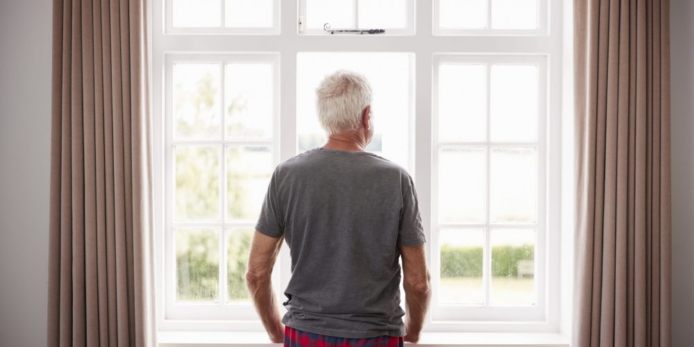 Rear View Of Senior Man In Pajamas Looking Out Of Bedroom Window